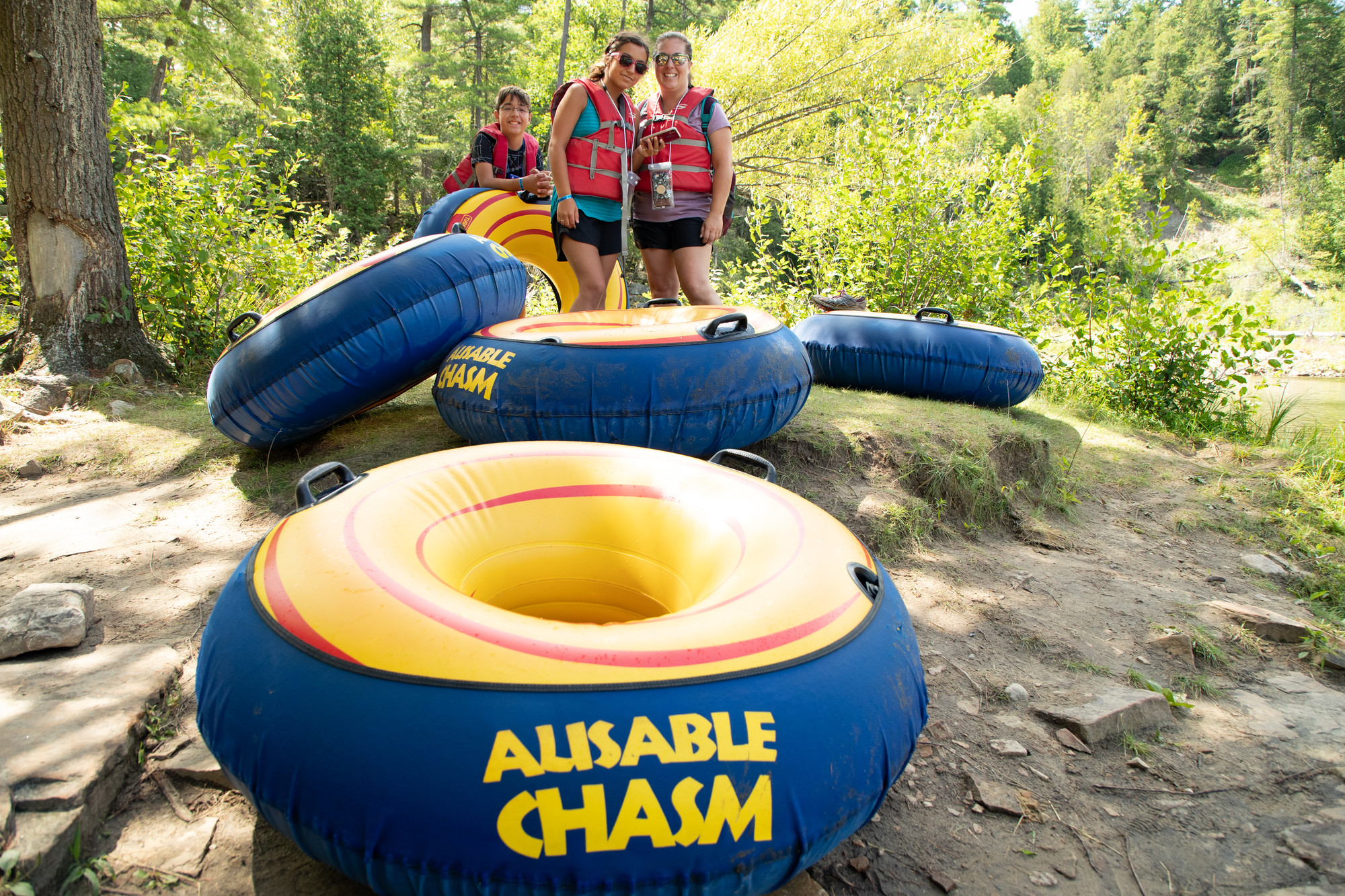 Two women and a child in life jackets pose next to inner tubes on a sunny day.