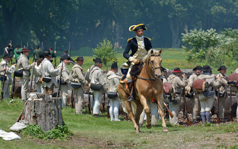 A costumed American Revolution reenactor rides a light brown horse past uniformed soldiers.