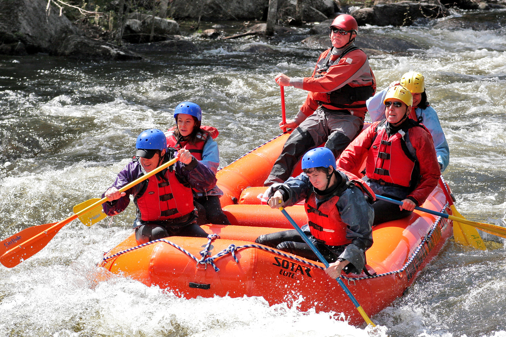 Six adults paddle in an inflated raft on a river busy with rapids.