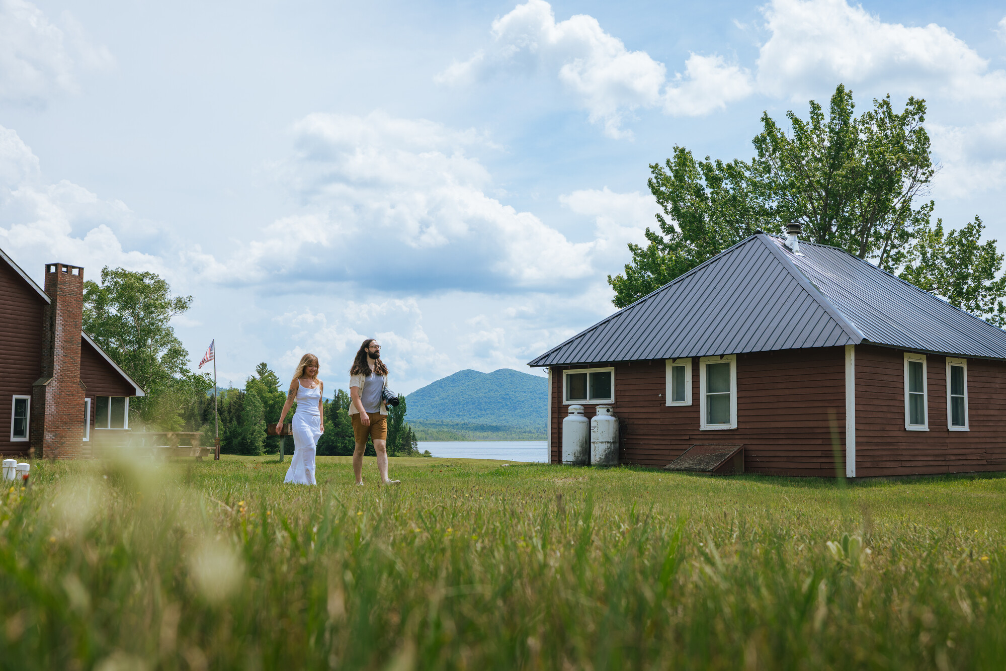 Two women walk toward a small square, one-story cabin with a lake and mountain in the distance.