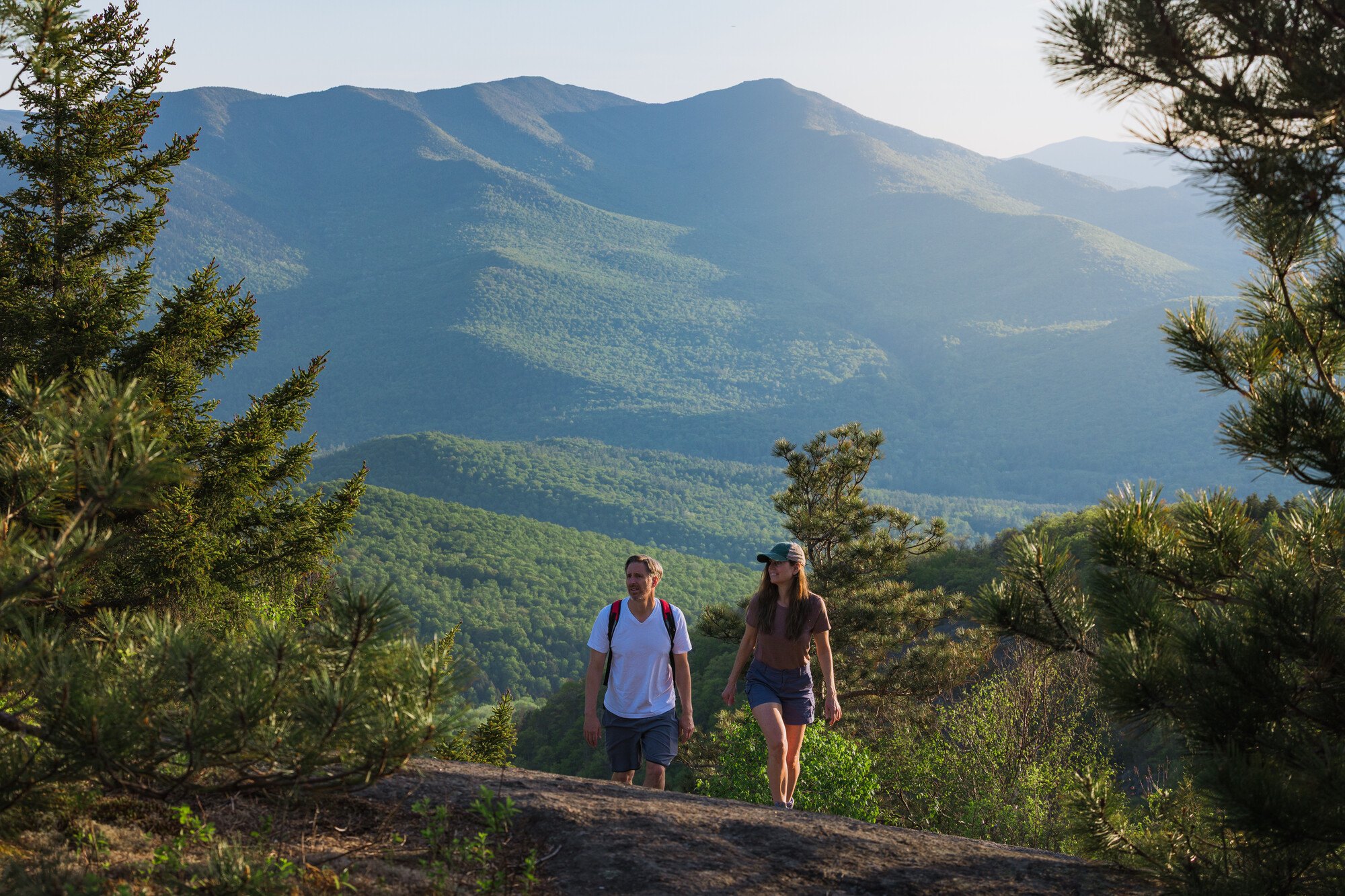Two adults ascend a rock while a large, forested mountain dominates the backdrop.