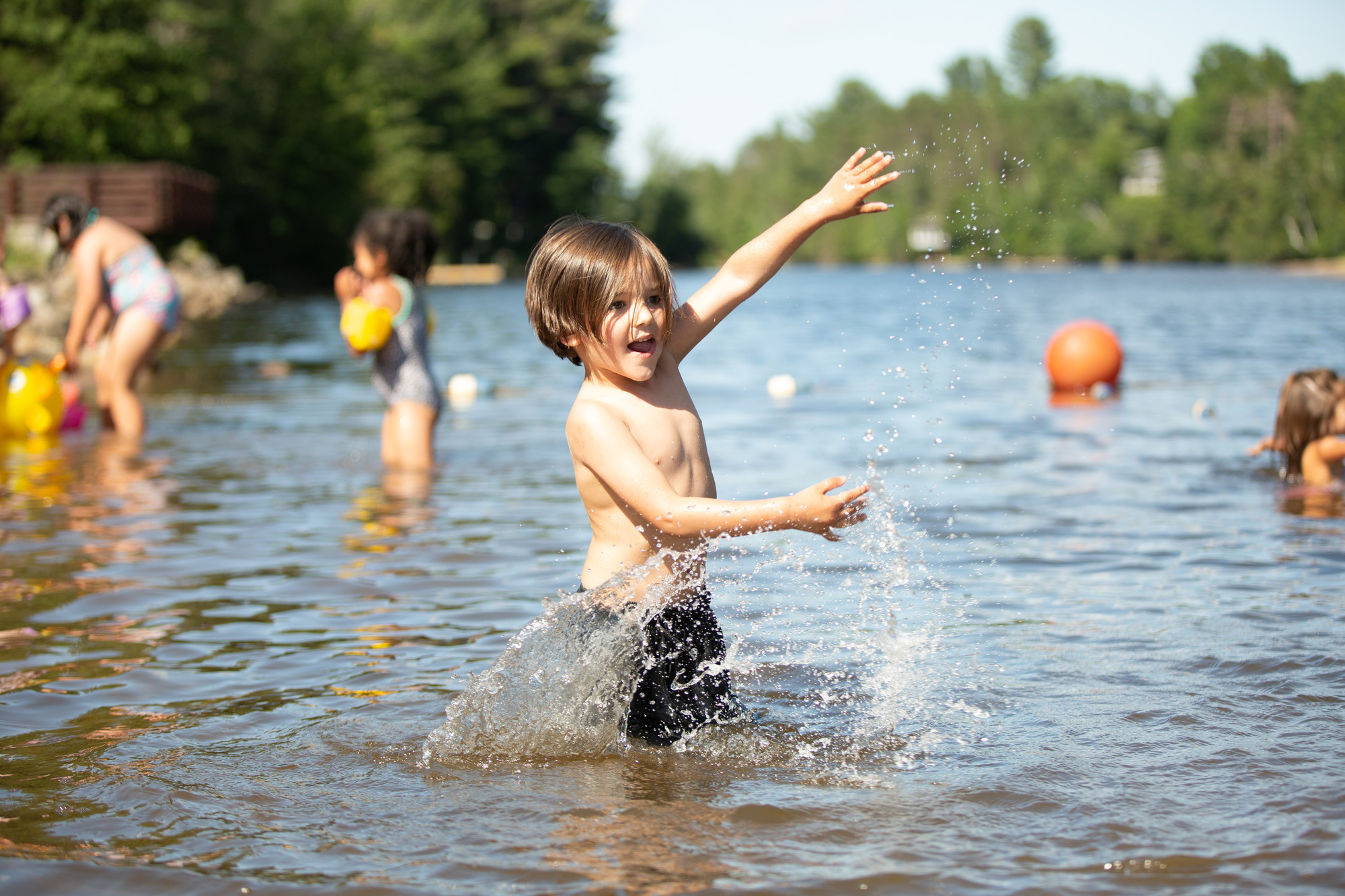 Small children splash and play in shallow water at a tree-lined beach.