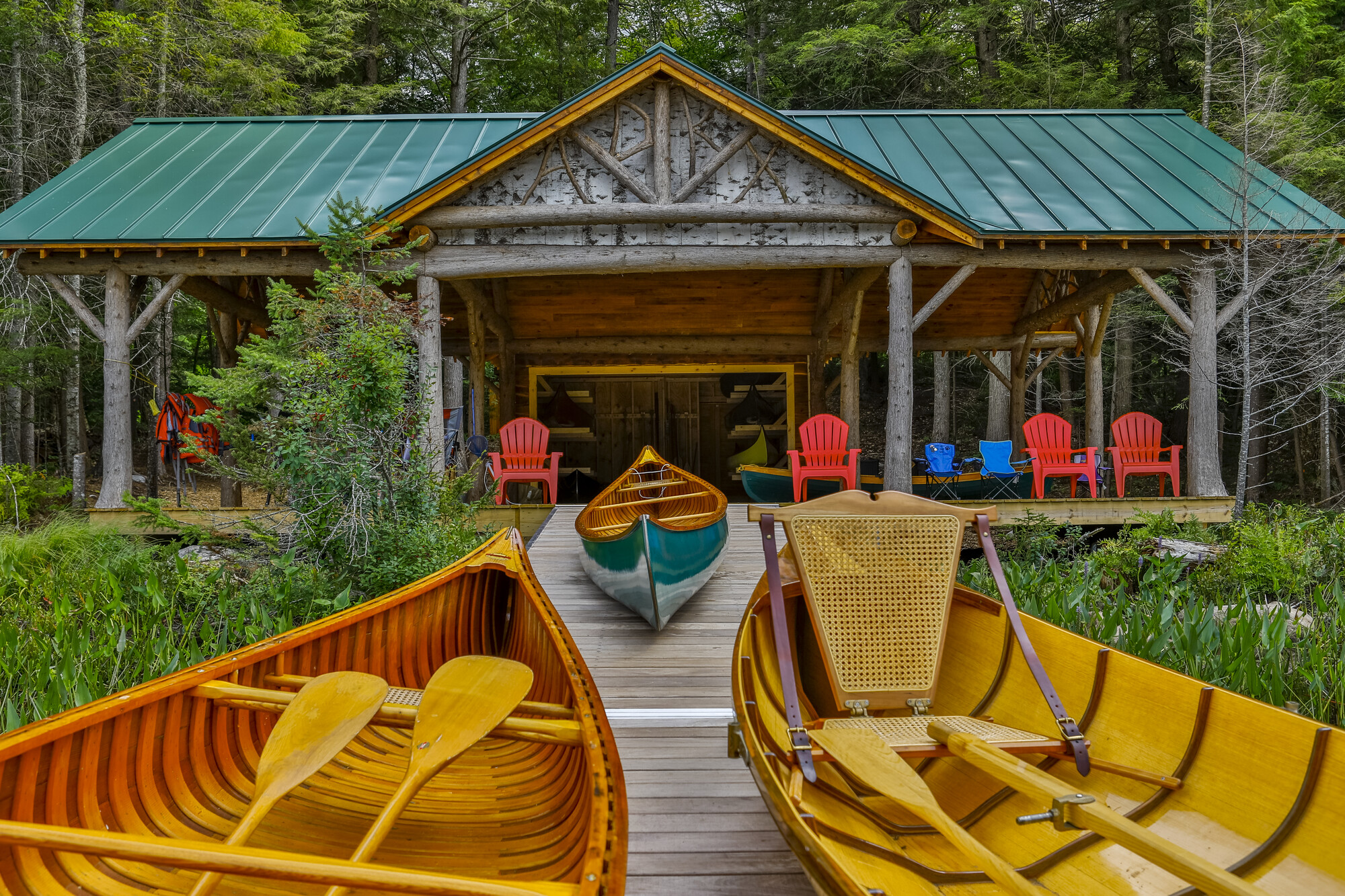 Three wooden guideboats rest on a dock in front of a rustic Adirondack boathouse.