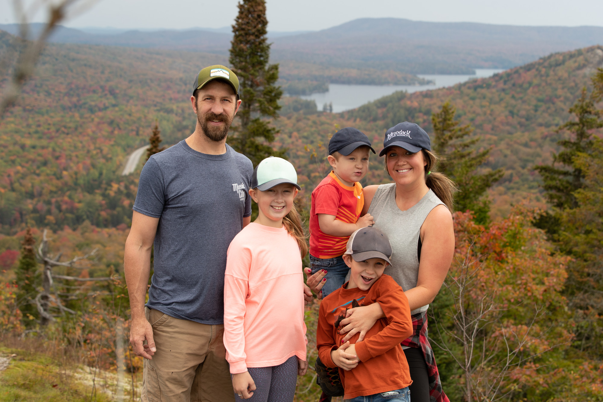 A man and woman and their three children smile for a picture on top of a mountain in fall. 