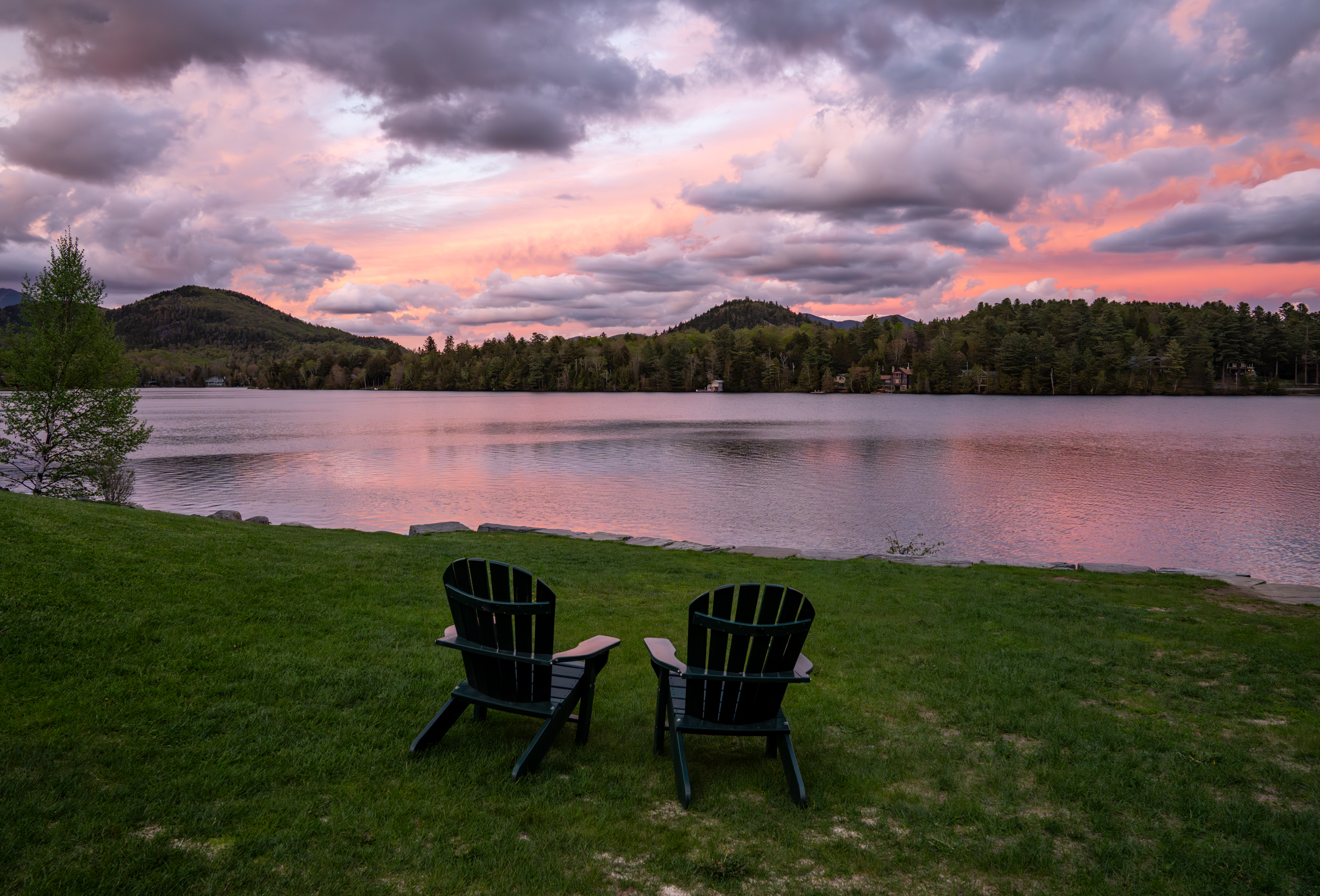 Two Adirondack chairs in front of a lake with a purple and pink sunset overhead.