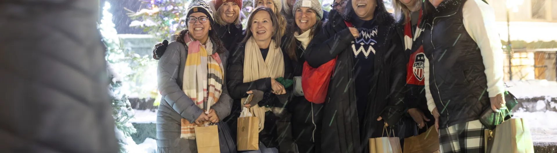 A group of ten women in winter outdoor clothing smile for a photo in front of a lit Christmas tree, holding shopping bags.