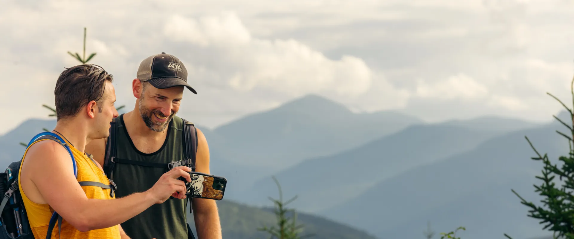 Two men look at a phone on the top of a mountain with mountains in the background. 
