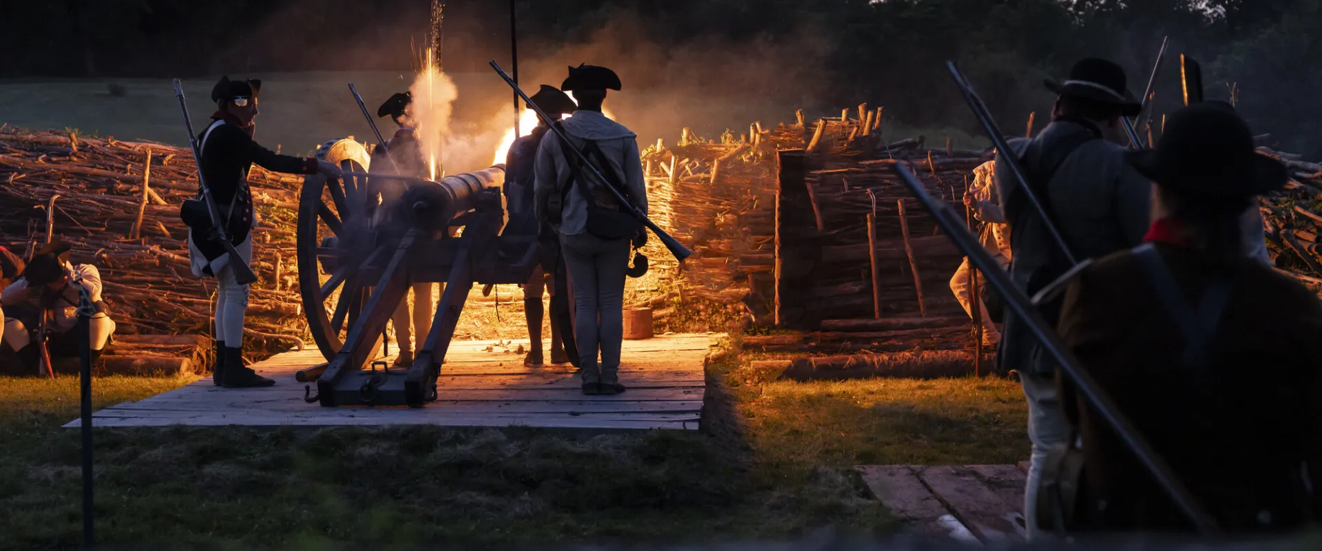 Costumed battle reenactors stand before a cannon lit by the glow of a fire.