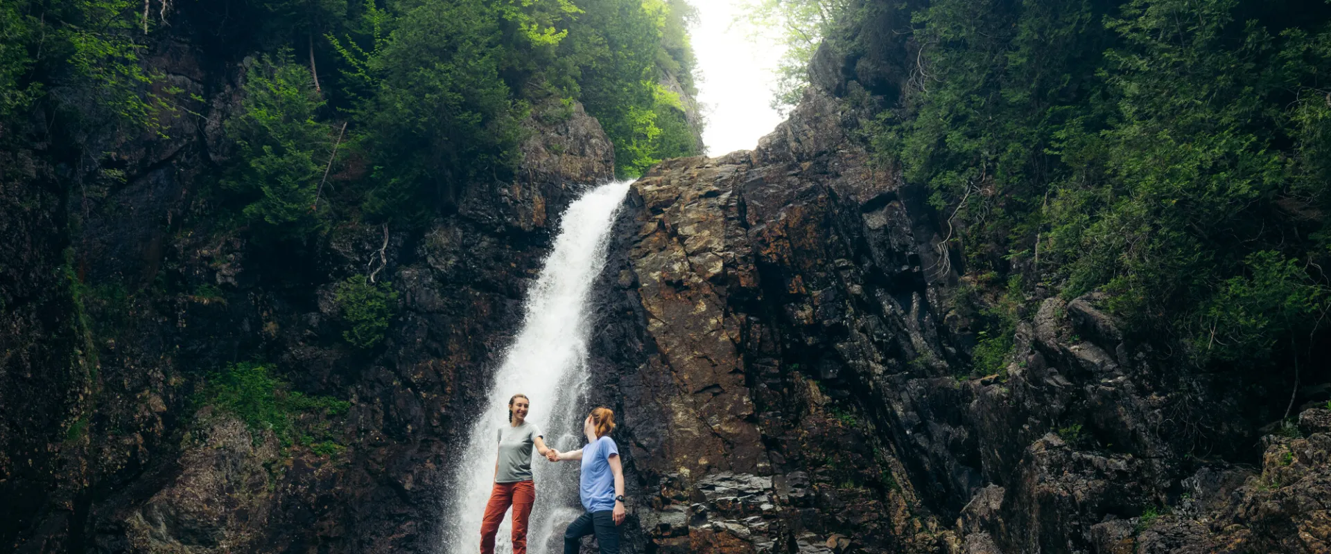 Two adults stand on rocks below a waterfall which tumbles between high, forested riverbanks.