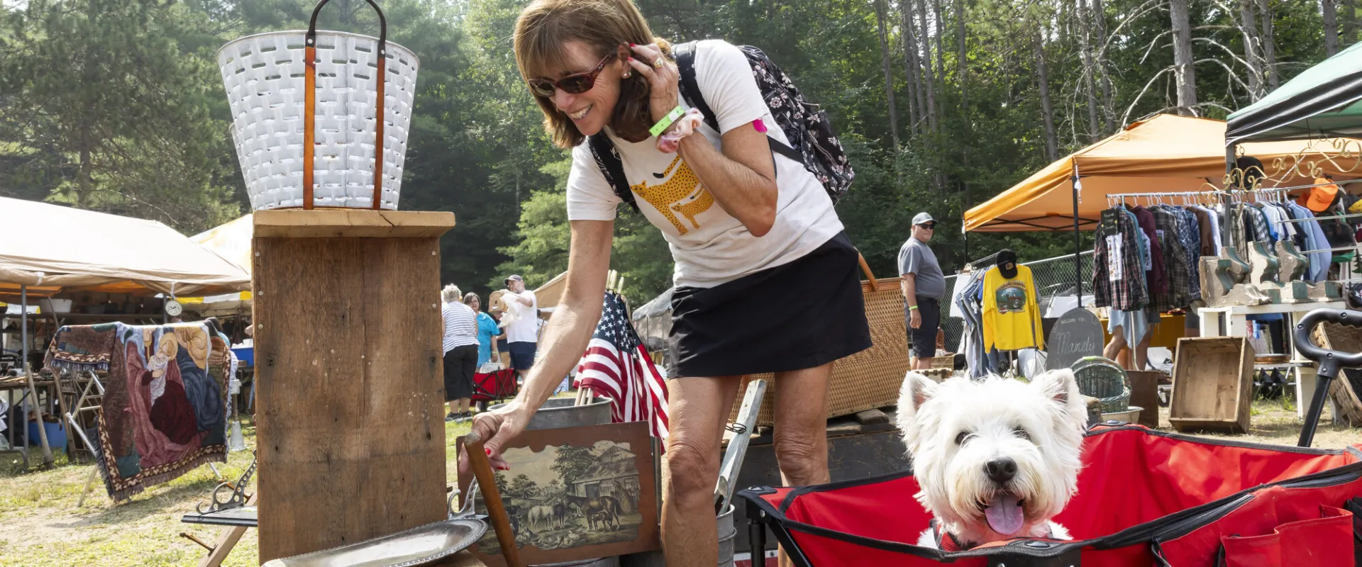 A small white dog looks cheerfully out from a red cart while his person browses at an antique show.