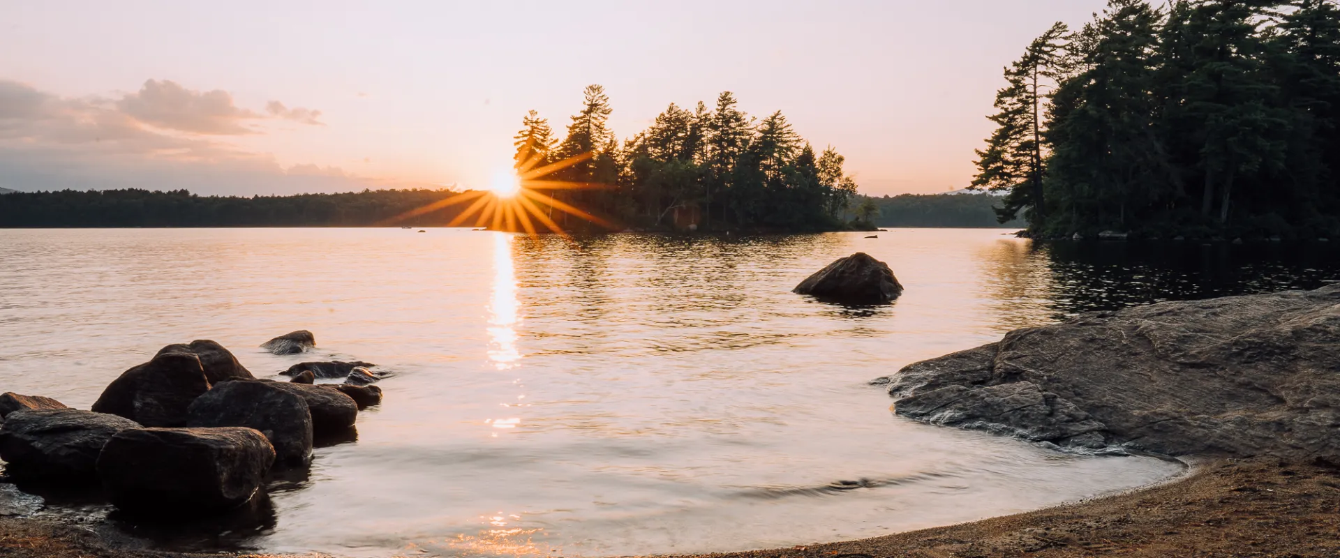 The sun sets behind a small, wooded island on a lake. The water reflects the colorful sky in front of a sandy beach.