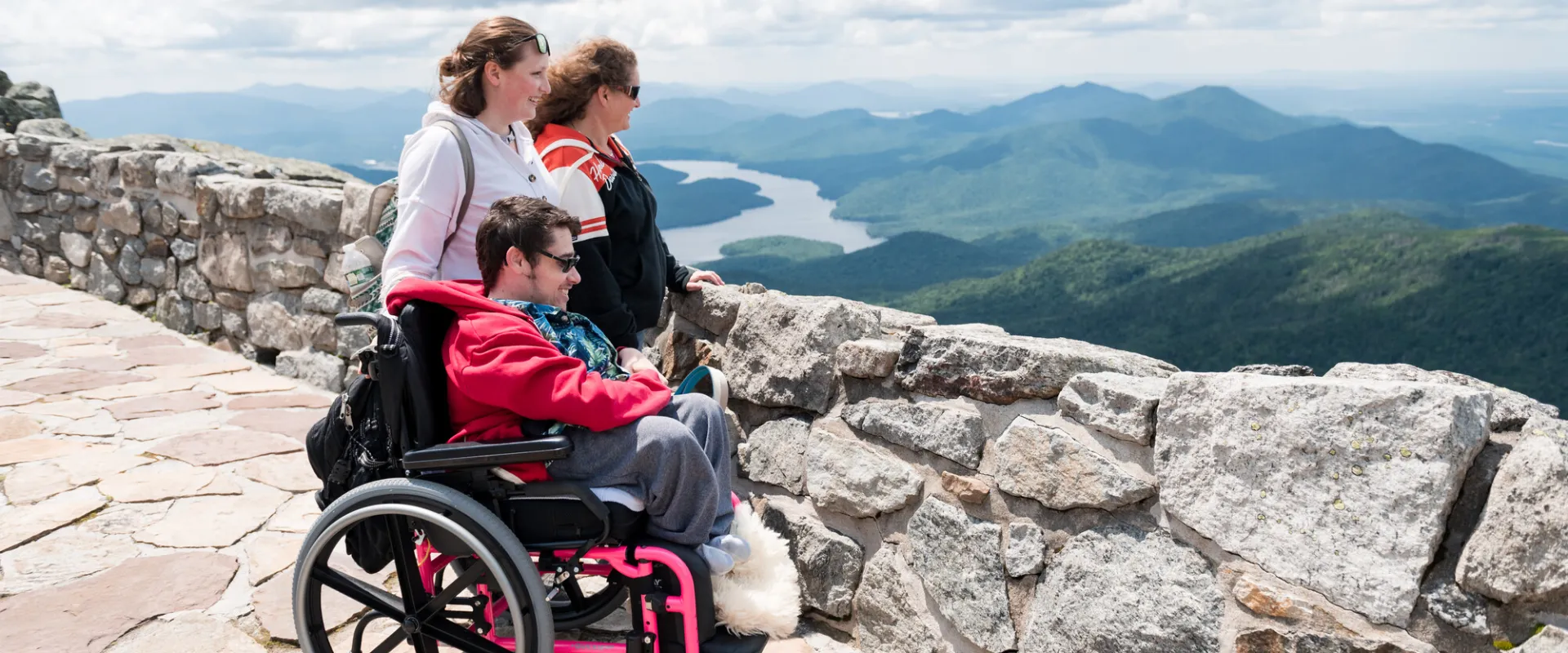A man in a wheelchair and two women stand on a stone trail on a mountain. 