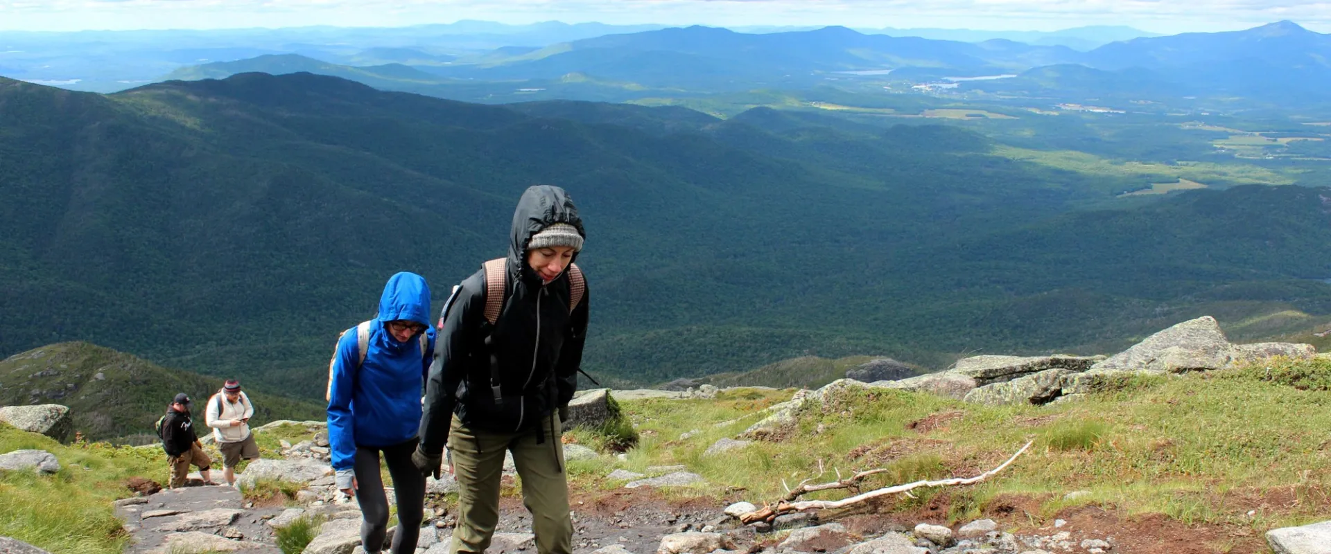 Two women climb up a mountain overlooking a valley and distant mountain ranges. 