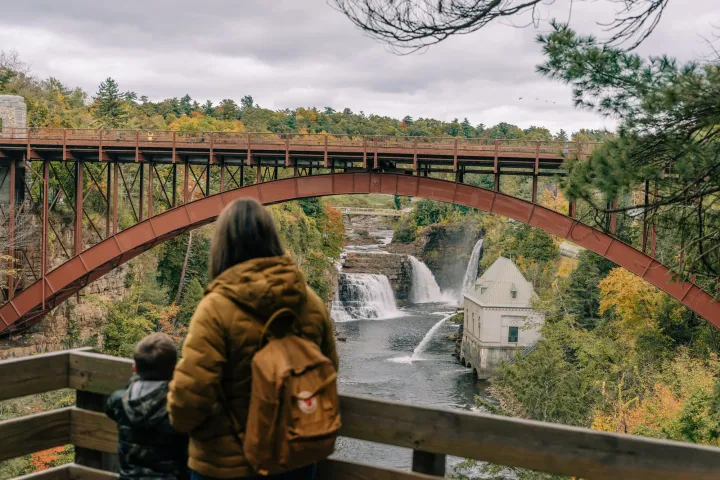 A woman and her young son look out at a bridge and mill with a waterfall in the background. the scene is surrounded by fall foliage. 