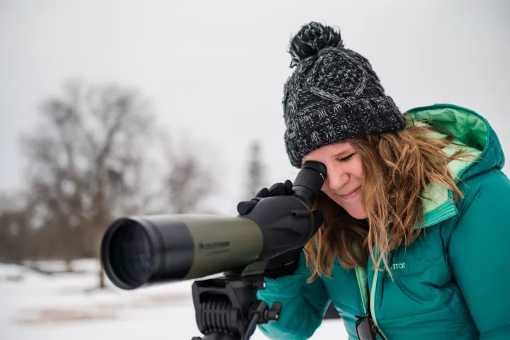 A woman in a teal jacket and beanie takes a picture through a long-lens camera. 