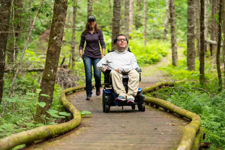 A man in a motorized wheelchair rides along a wooden path with a woman walking behind him. 