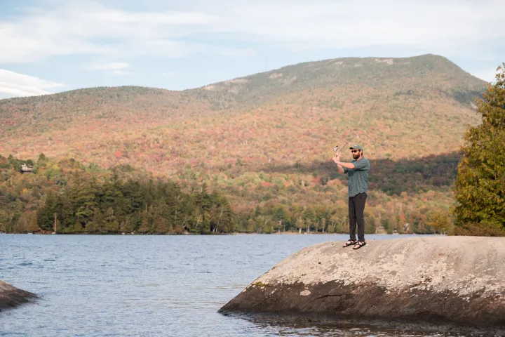 A man fishing on a rock peninsula in front of fall foliage