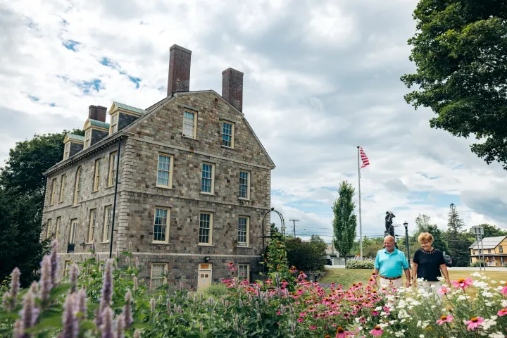 A man and woman walk alongside a colonial-style brick home. 