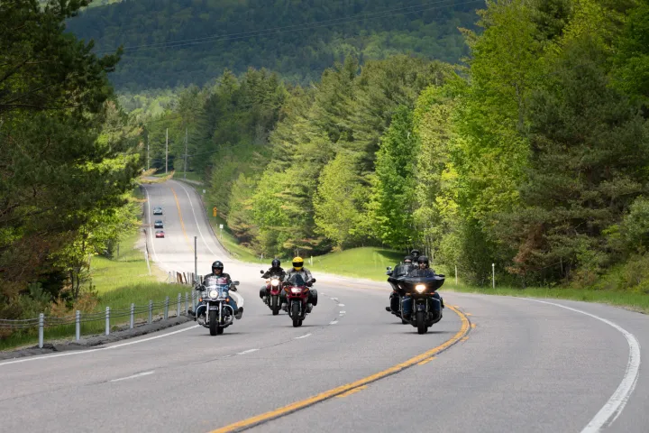 A group of bikers ride along a road with mountains in the background. 