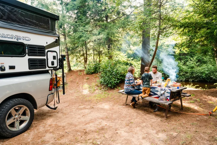 Two adults and a child sit at a campsite picnic table. A grill smokes behind them and a camper is viewed to the left.