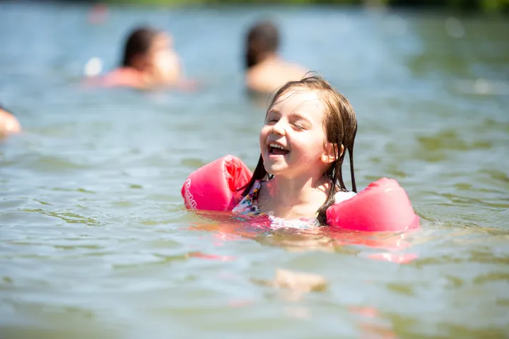 A little girl laughs while swimming in shallow water, wearing pinkish-red water wings for safety.