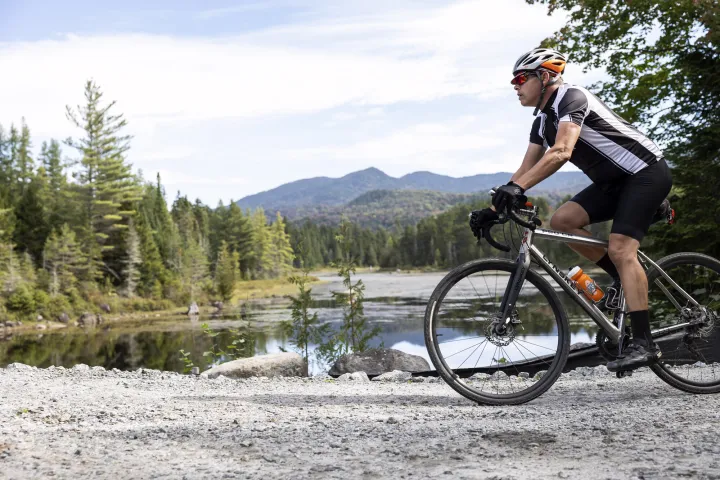 A cyclist rides a mountain bike past a pond and large mountain range in the Adirondacks.