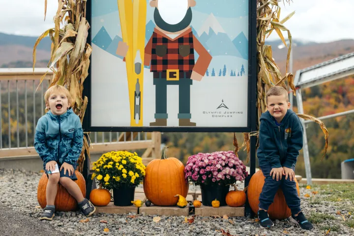 Two small boys grin while sitting on pumpkins at a harvest festival.