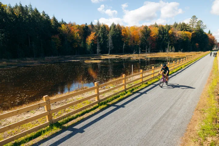 A lone cyclist rides along a wooden rail fence on a smooth gravel trail on a sunny fall day.