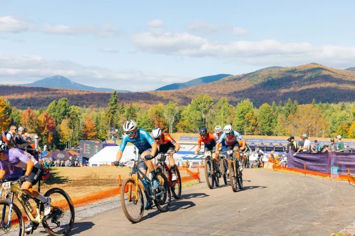 Mountain bike riders ride up a gravel hill in a competition surrounded by foliage-covered mountains and spectators.