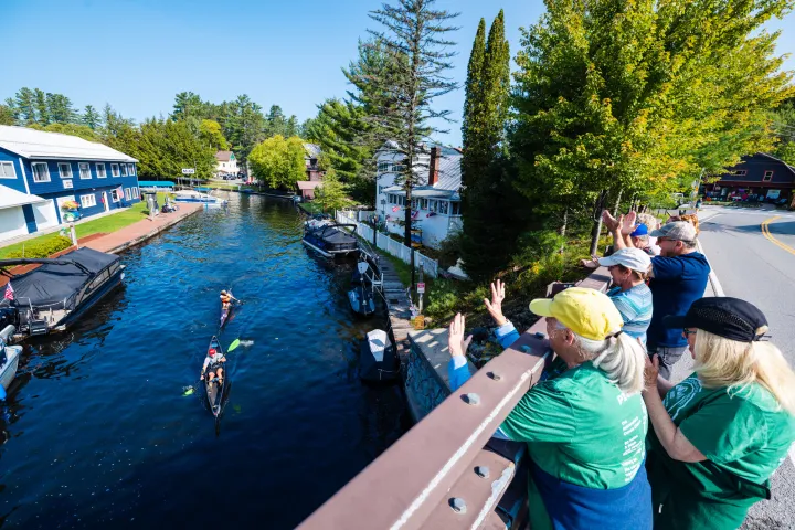 Spectators cheer on paddlers passing under a small bridge on a sunny day.
