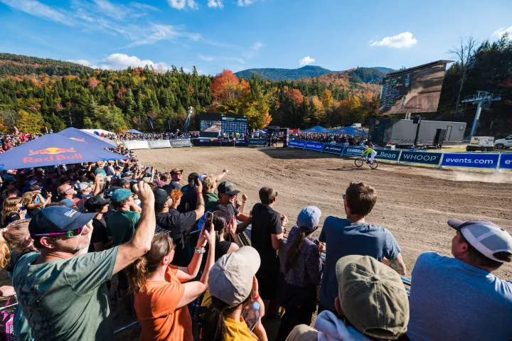 A crowd watches the finish of a mountain bike race with fall foliage on the mountains in the distance.
