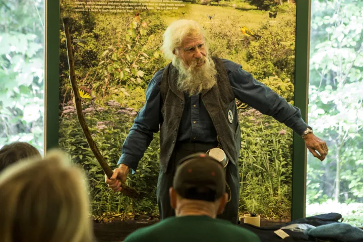 A bearded, elderly Adirondack guide speaks before a scenic, green backdrop at an event.