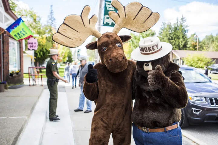 Two costumed characters&mdash;a moose and Smokey&mdash;give the thumbs up to the camera on a sunny day on a small town sidewalk.