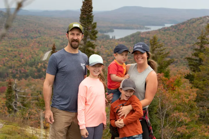 A man and woman and their three children smile for a picture on top of a mountain in fall. 