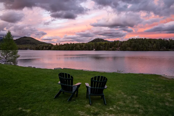 Two Adirondack chairs in front of a lake with a purple and pink sunset overhead.