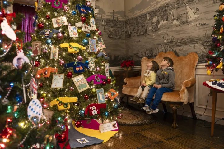 A Christmas tree with a variety of colorful ornaments. Two children sit in chairs in the background.