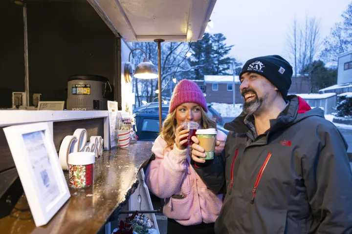 A woman and a man drink cups of hot chocolate at an outdoor stand at a winter event.