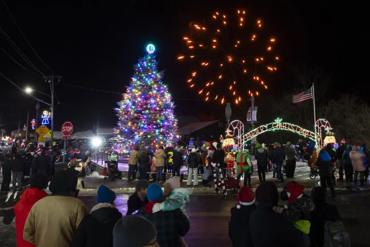 Fireworks explode over a large lit Christmas tree and a crowd at a Christmas festival.