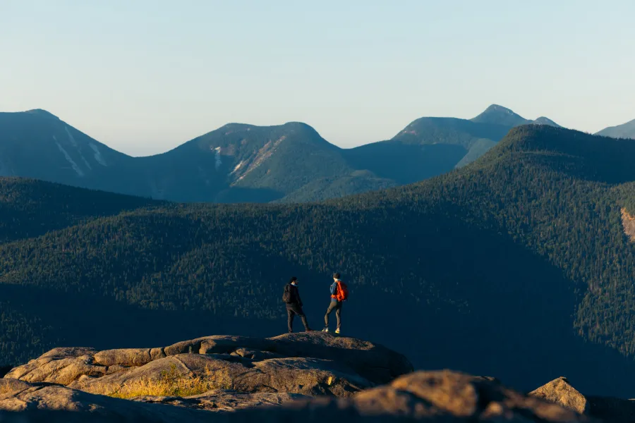 Two people on a summit looking out at big mountains during sunrise