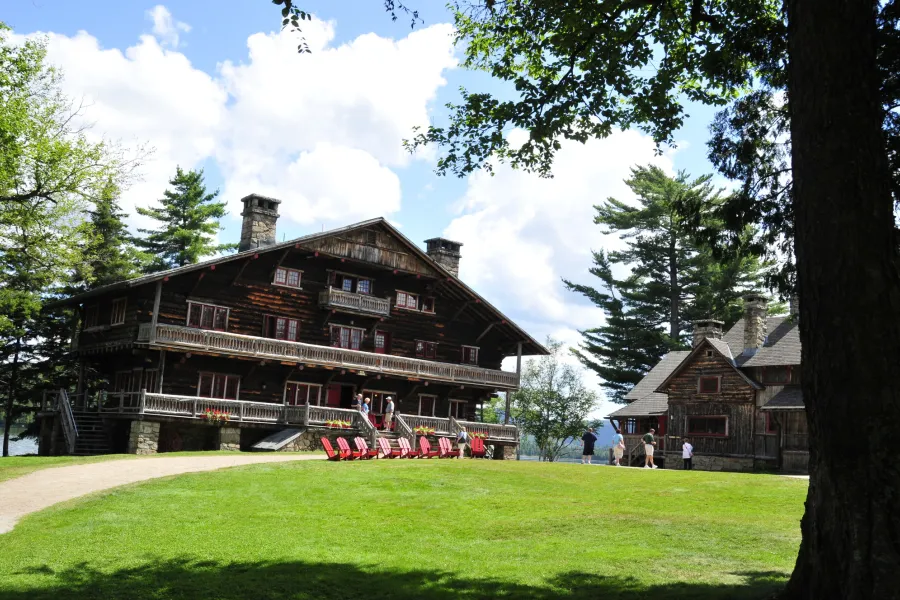 An Adirondack Great Camp lodge sits on a trimmed lawn with red Adirondack chairs in front of it. 