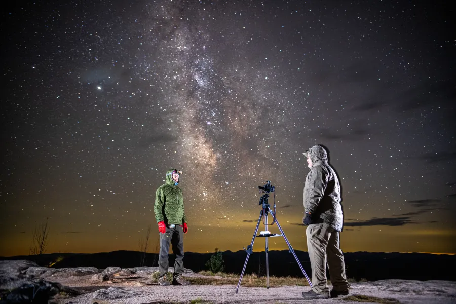 Two men stand on top of a mountain at night to stargaze with a camera.