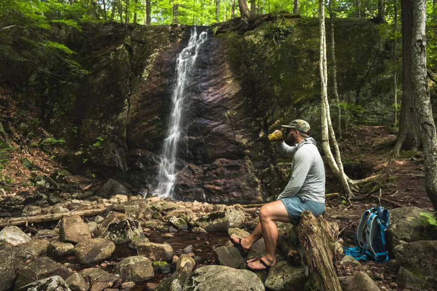 A hiker drinking water in front of a ribbony waterfall