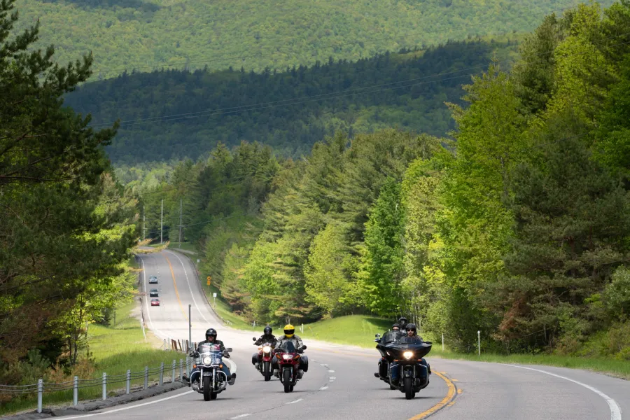 A group of bikers ride along a road with mountains in the background. 