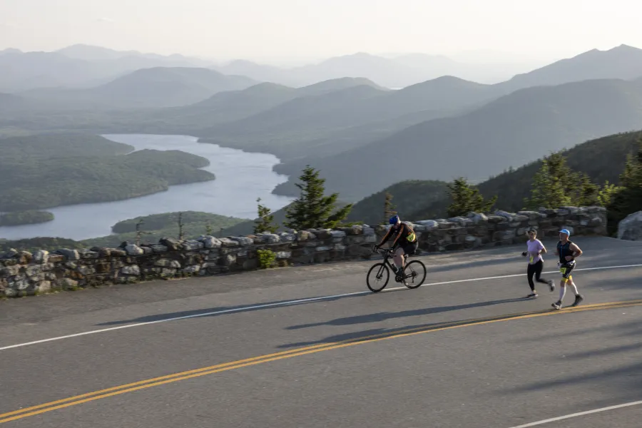 Runners and a man on a bike climb a mountain road overlooking a lake and mountains below. 