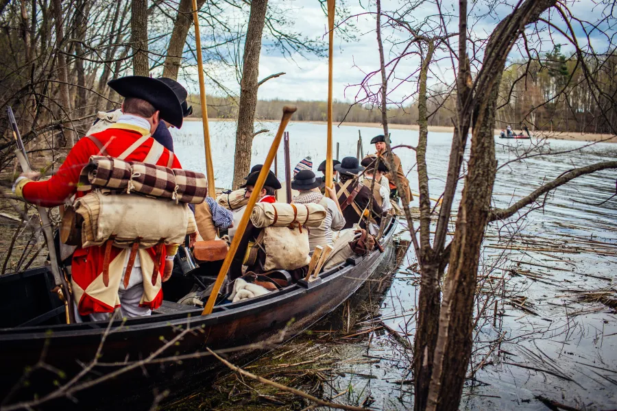 A boat containing costumed American Revolution reenactors is tucked into the reeds of a lake.