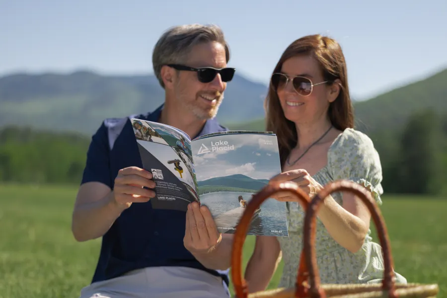 A man and woman smile while looking at a tourism visitor guide on a sunny day with a scenic mountain background.