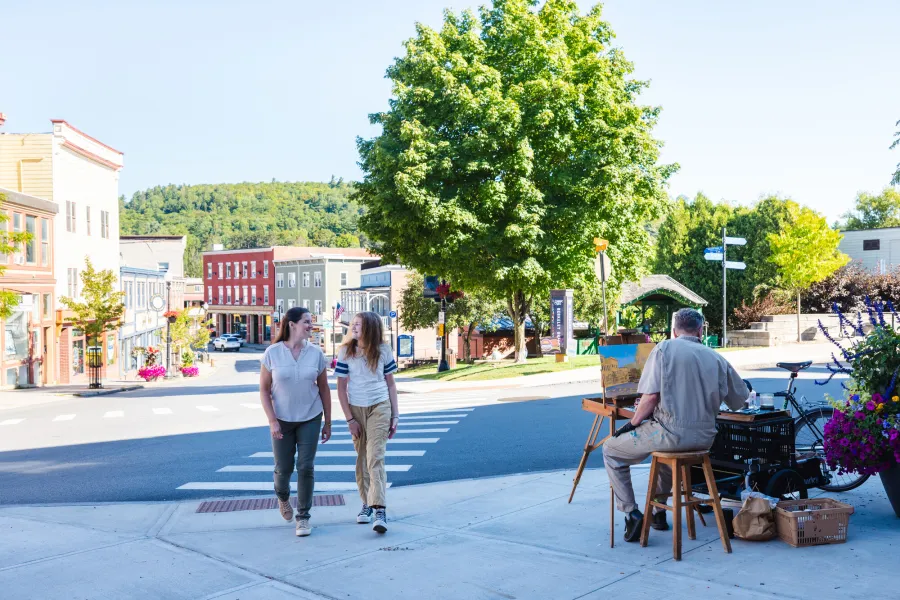 Two women walk on a sidewalk past an artist working at an easel on a sunny summer day.