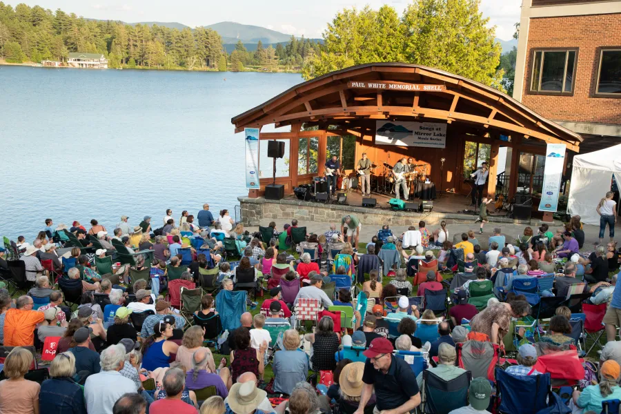 A hill covered with people watching an outdoor concert at a lakeside bandshell on a summer evening.