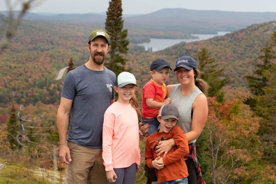 A man and woman and their three children smile for a picture on top of a mountain in fall. 