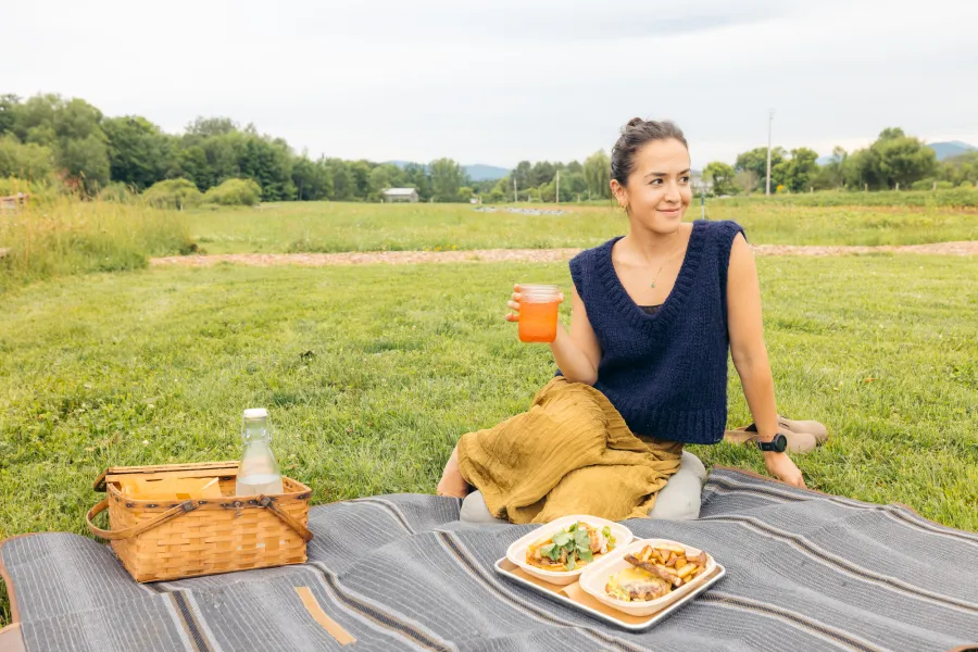 A woman holds a cup of beer while sitting on a picnic blanket on a field. A picnic basket and plate of food are next to her.